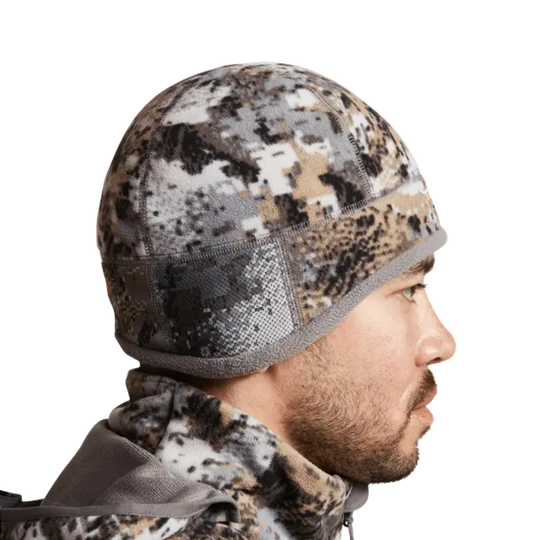 Person wearing a camouflage beanie and jacket on a white background