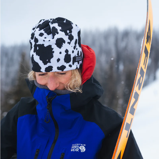 Person wearing a blue jacket and black and white patterned hat with a red hood, holding a ski.