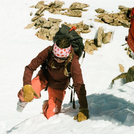 Woman wearing maroon jacket and peach snow pants on snowy rocky terrain. Wearing pink strawberry headband