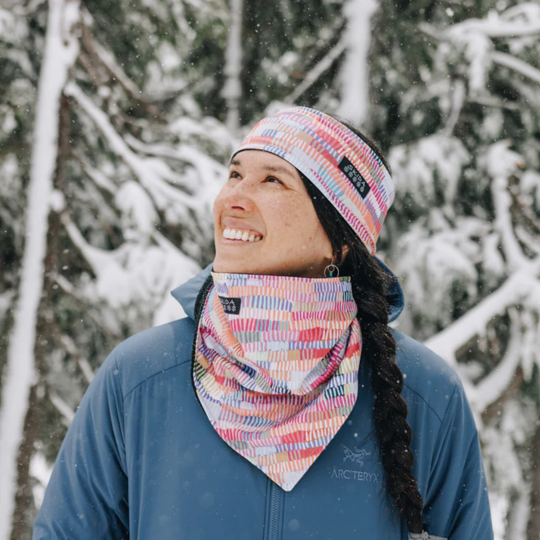Woman wearing Multi color Penny Lane headband with skida logo and matching neck warmer. Blue jacket on a snowy pine tree background 