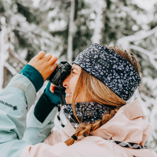 Woman wearing Black and white flower print headband and matching neck warmer. On a snowy pine tree background looking through a camera.