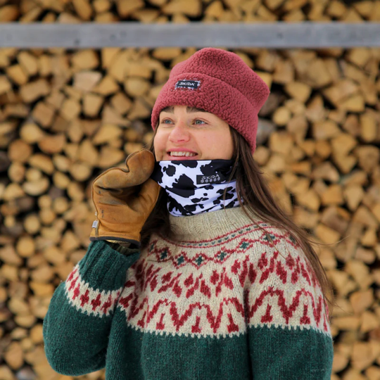 Person wearing winter clothing with a patterned sweater, beanie, and gloves against a background of stacked firewood.
