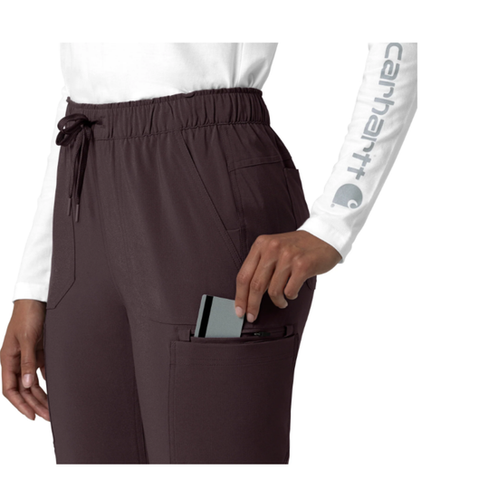 A close-up of a person wearing dark brown drawstring scrub pants with a side pocket holding a phone, paired with a white top. The person's right arm is visible wearing a white Carhartt long-sleeve shirt, and the Carhartt logo is printed on the sleeve.
