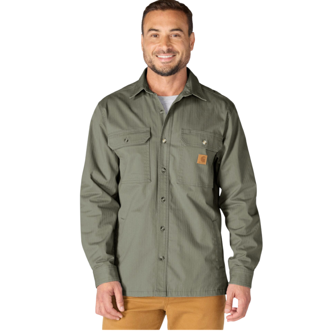 A man wearing a long-sleeve button-up work shirt in olive green with two chest pockets and a small orange Carhartt logo patch, paired with tan pants, against a plain white background.