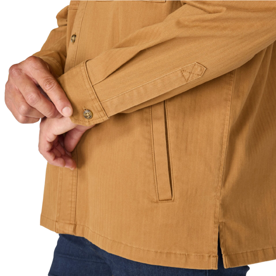 Close-up of a person wearing a tan long-sleeve button-front shirt jacket, showing the sleeve cuff with metal buttons and a vertical welt pocket, against a plain white background.