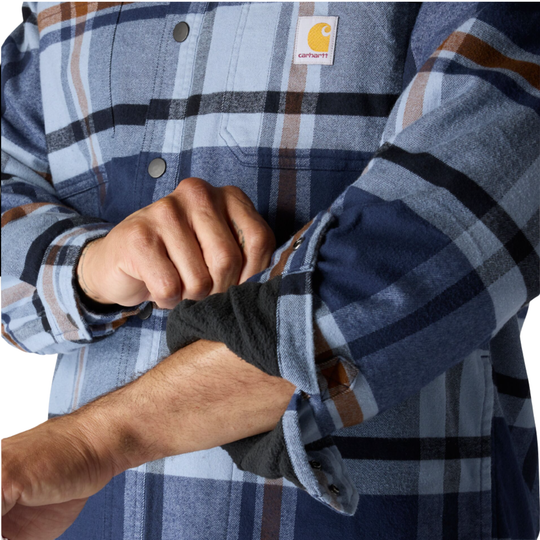 Close-up of a person wearing a blue and navy plaid Carhartt flannel shirt, rolling up the sleeve to reveal a dark fleece lining. The Carhartt logo patch is visible on the chest.