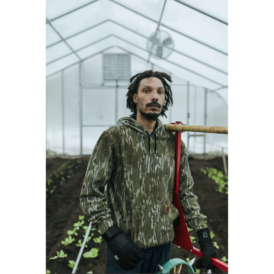 A man wearing a camouflage hoodie and black gloves stands inside a greenhouse holding a red wheelbarrow.