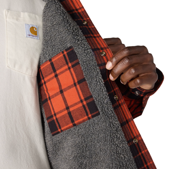 Close-up of a person wearing a Carhartt flannel shirt with a gray sherpa lining. The shirt features a red and black plaid pattern, a matching plaid chest pocket, and a visible Carhartt logo patch on the left chest pocket.