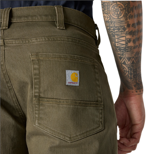 Close-up of a person wearing olive green Carhartt work pants, showing the back pocket with the signature Carhartt logo patch and part of the model's tattooed arm against a white background.