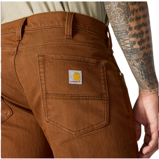 Close-up of a person wearing brown Carhartt work jeans, showing the back pocket with the Carhartt logo patch and part of the tattooed arm.