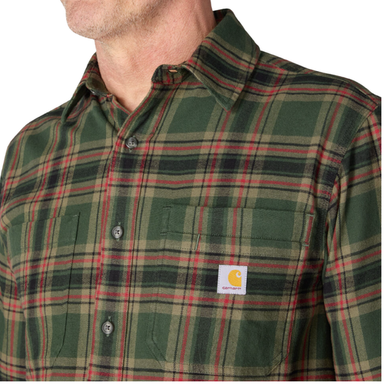 A close-up of a man wearing a Carhartt plaid button-down shirt in forest green with red and black stripes, featuring the signature Carhartt brand patch on the chest pocket.