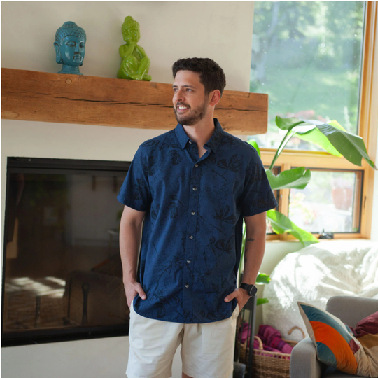 A man wearing a navy blue short-sleeve button-down shirt with a subtle tonal floral pattern and off-white shorts, standing in a brightly lit living room.