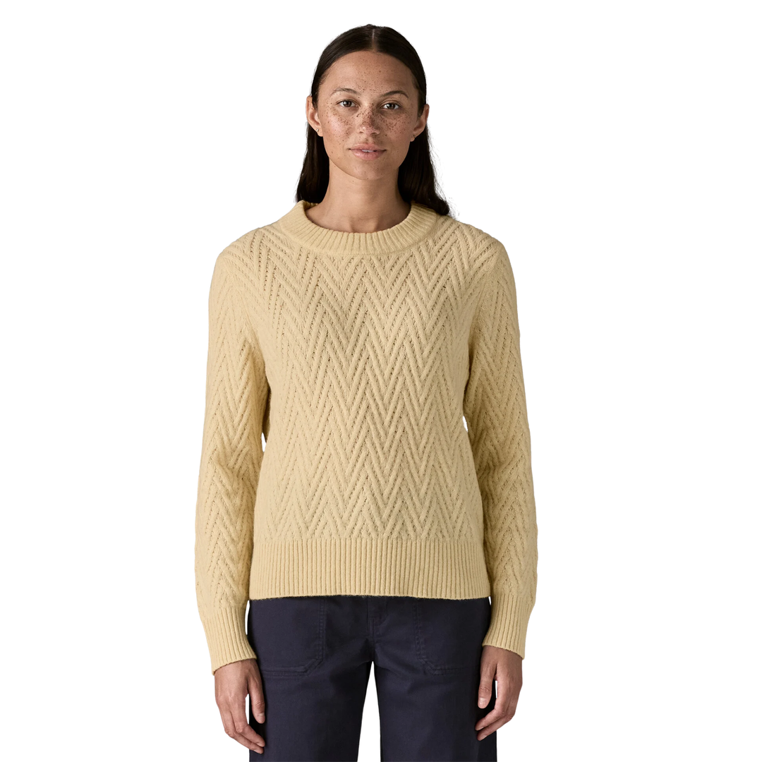 A woman modeling a cream-colored cable-knit crewneck sweater with a textured chevron pattern, paired with dark navy pants, against a plain white background.