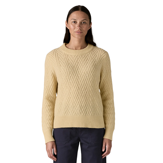 A woman modeling a cream-colored cable-knit crewneck sweater with a textured chevron pattern, paired with dark navy pants, against a plain white background.