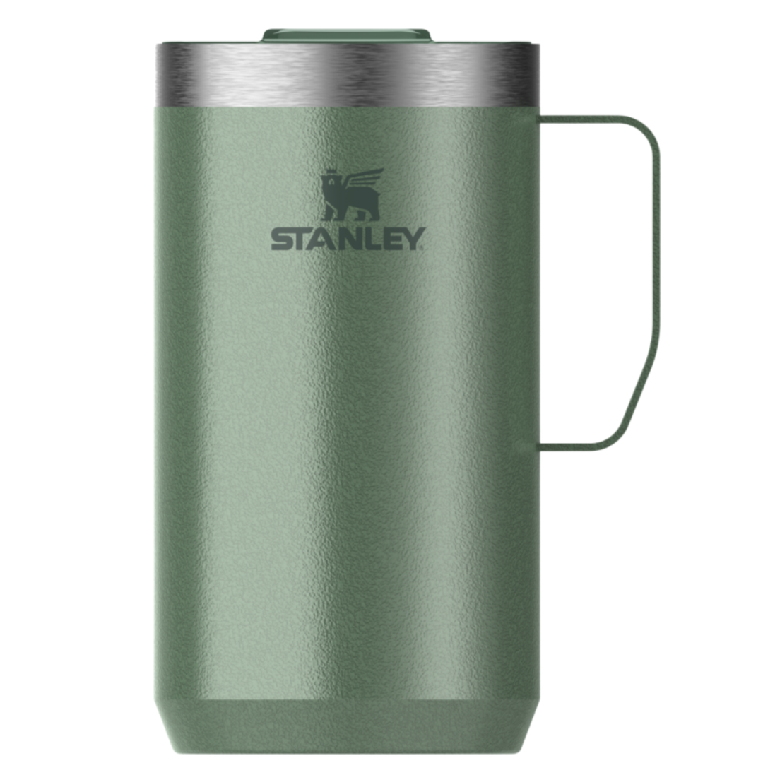 A Stanley travel mug in a textured sage green finish, featuring a stainless steel rim, a dark green handle, and the Stanley logo printed on the front, set against a plain white background.