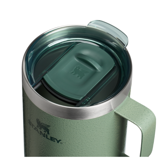 Close-up of a Stanley insulated mug with a textured sage green body, stainless steel rim, and matching green lid with a clear flip-up straw cover, shown on a white background.