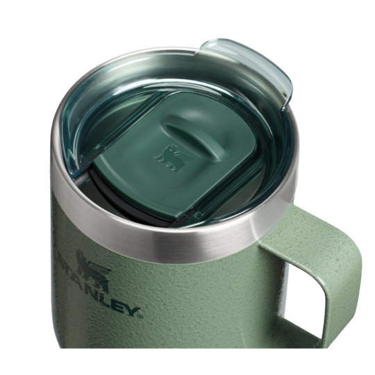 A close-up of a Stanley insulated mug with a textured sage green body, a stainless steel rim, and a translucent dark green lid with a clear flip-up spout.