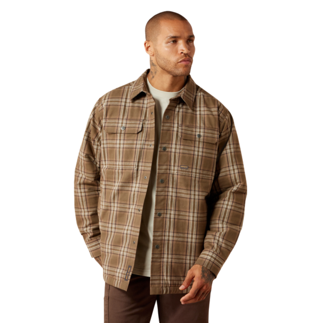 A man wearing a brown and beige plaid button-up overshirt over a light gray t-shirt and brown pants, photographed against a solid white background.