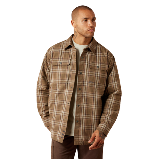 A man wearing a brown and beige plaid button-up overshirt over a light gray t-shirt and brown pants, photographed against a solid white background.