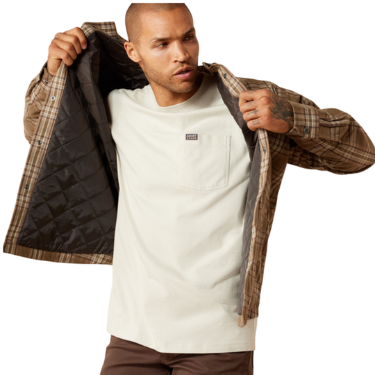A man wearing a brown and beige plaid flannel shirt jacket with a black quilted lining, layered over a white t-shirt and brown pants, holding the jacket open against a white background.