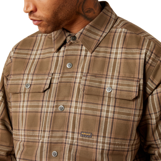 A close-up of a man wearing a brown plaid button-up shirt with silver buttons and chest pockets. The shirt features a subtle check pattern in brown, beige, and white, with a small 'AHAT' brand label near the lower right chest.