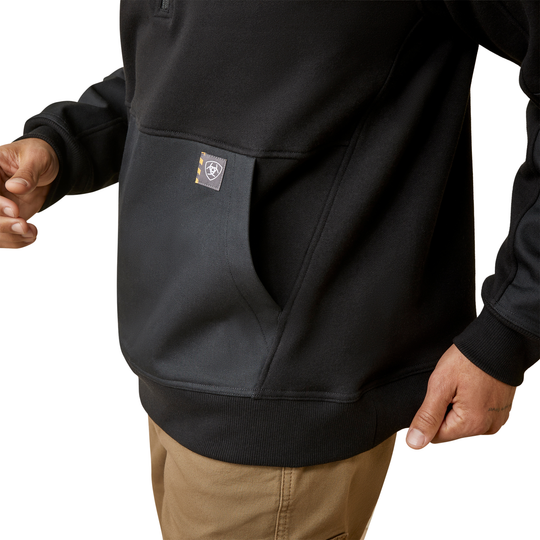 A close-up of a person wearing a black pullover hoodie with a kangaroo pocket and a small square logo patch on the pocket, paired with tan pants against a white background.