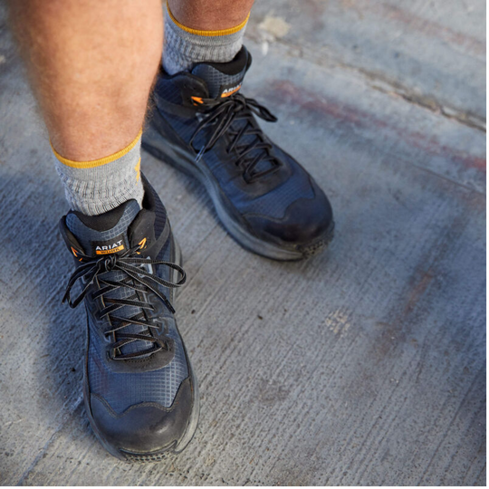 A close-up of a person wearing black and dark gray high-top work boots with gray socks, standing on a concrete surface. The brand name 'ARIZ' is visible on the tongue of the boots.