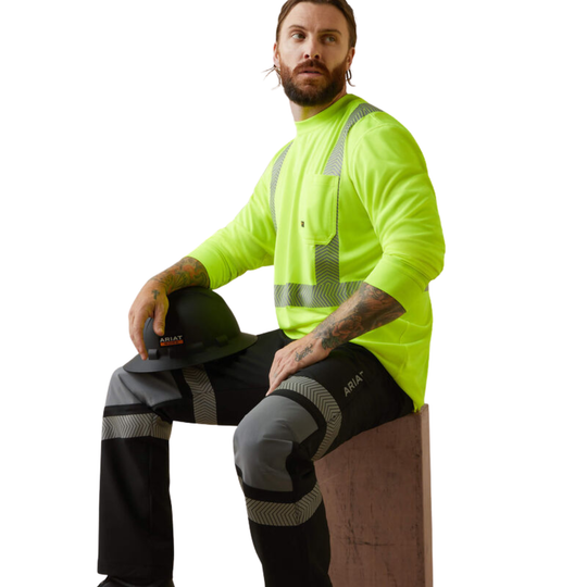 A man wearing a high-visibility neon yellow long-sleeve work shirt with reflective straps, black high-visibility trousers with gray reflective bands, and a black hard hat, seated on a wooden block against a white background.