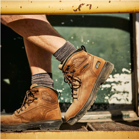 A person wearing brown suede work boots with gray knit socks, sitting on a rusty yellow metal structure.
