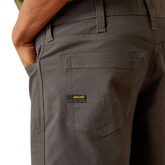Close-up of a person wearing dark gray Ariat work pants, showing the back pocket with a small yellow ARIAT logo patch.