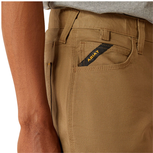 Close-up of a person wearing tan work pants, showing the back pocket with a black Ariat brand tab and part of the gray shirt at the waist.