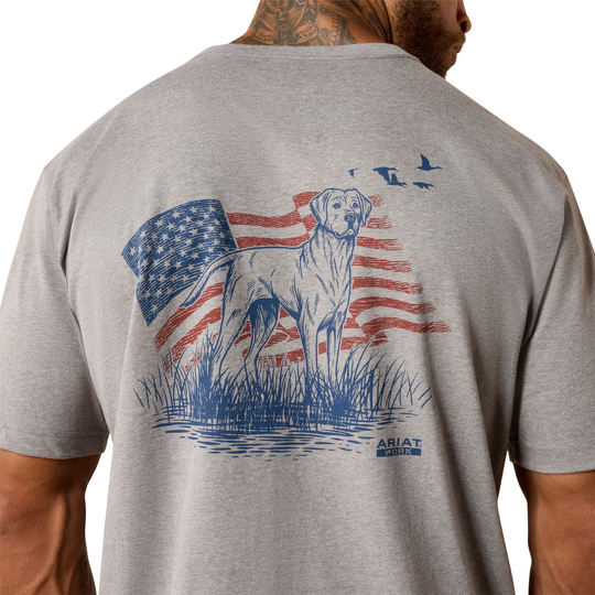 A man wearing a heather gray t-shirt, viewed from the back. The shirt features a graphic print of a white dog standing in front of a distressed American flag, with blue birds flying above and a blue 'ARIAT' logo at the bottom right.