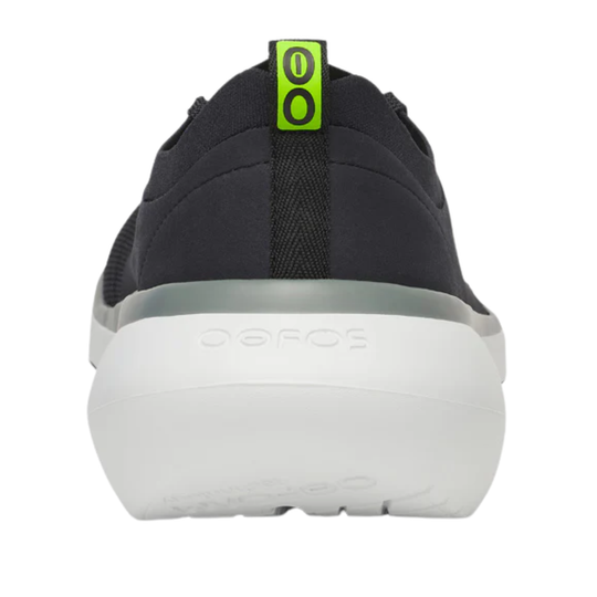 A close-up rear view of a black Oofos sneaker showing the thick white sole and a bright neon green pull tab with black 'OO' lettering.