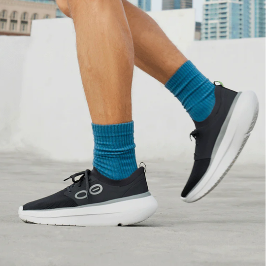 A person wearing black athletic sneakers with thick white soles and bright blue ribbed socks, photographed outdoors on a light concrete surface with buildings blurred in the background.