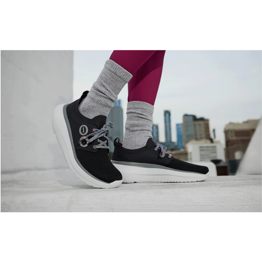 A person wearing black athletic sneakers with gray laces and white soles, paired with gray socks and magenta leggings, standing on a rooftop with a blurred city skyline in the background.