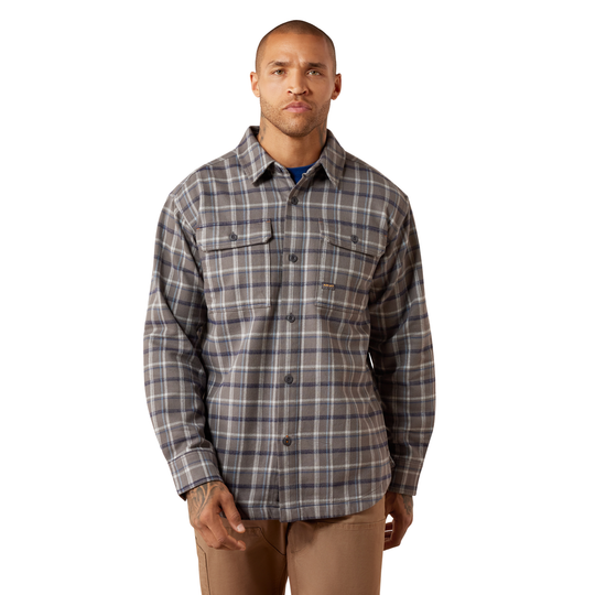 A man wearing a long-sleeve button-up plaid shirt in gray, white, and navy, paired with tan pants, against a plain white background.