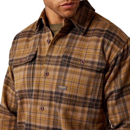 A close-up of a man wearing a brown and tan plaid button-up shirt jacket with a small visible Ariat logo patch on the chest pocket.