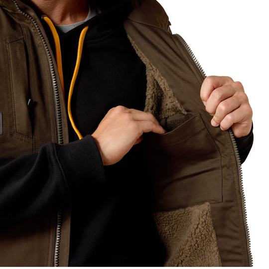 Close-up of a person wearing an olive brown utility jacket with a tan sherpa lining, opening a side pocket to show the interior. The person is also wearing a black hoodie with yellow drawstrings.