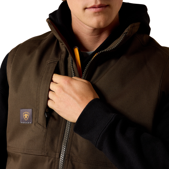 A close-up of a person wearing a dark brown hooded zip-front vest over a black hoodie. The vest has a small square chest patch with a yellow logo and a silver zipper running down the front.