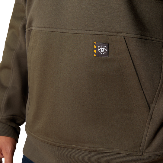 Close-up of an olive green pullover hoodie with a front kangaroo pocket and a small black brand patch on the pocket, worn by a model.