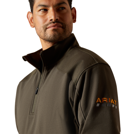 A man wearing a dark olive green half-zip pullover jacket with an orange and white ARIAT logo on the left sleeve, shown against a plain white background.