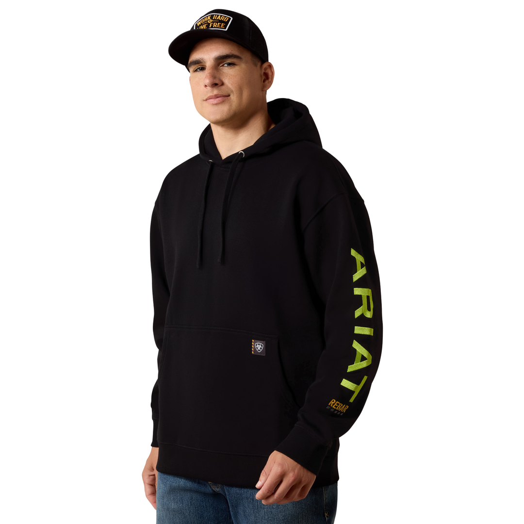 A man wearing a black Ariat pullover hoodie with a bright yellow-green vertical ARIAT logo on the left sleeve, paired with a black baseball cap and blue jeans, against a plain white background.