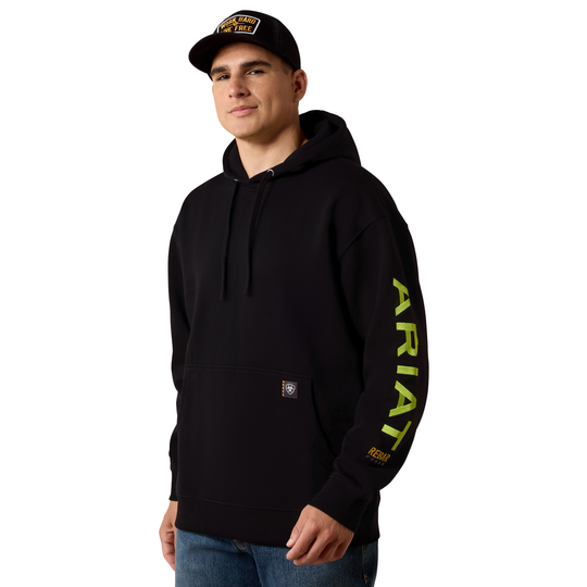 A man wearing a black Ariat pullover hoodie with a bright yellow-green vertical ARIAT logo on the left sleeve, paired with a black baseball cap and blue jeans, against a plain white background.