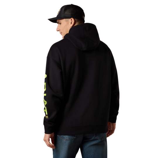 A man wearing a black pullover hoodie with a bright neon yellow-green vertical graphic print on the left sleeve, shown from a rear three-quarter view. He is also wearing a black trucker cap and blue jeans against a plain white background.