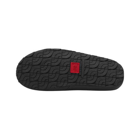 The black textured rubber outsole of a shoe, featuring a repeating wave-like tread pattern and a small red square logo patch in the center, shown against a plain white background.