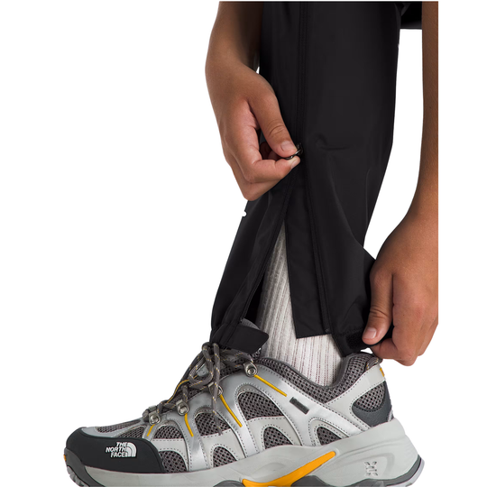 A close-up of a person's hands and lower legs as they pull up black pants over a white sock, wearing a gray and black The North Face hiking shoe with yellow accents.