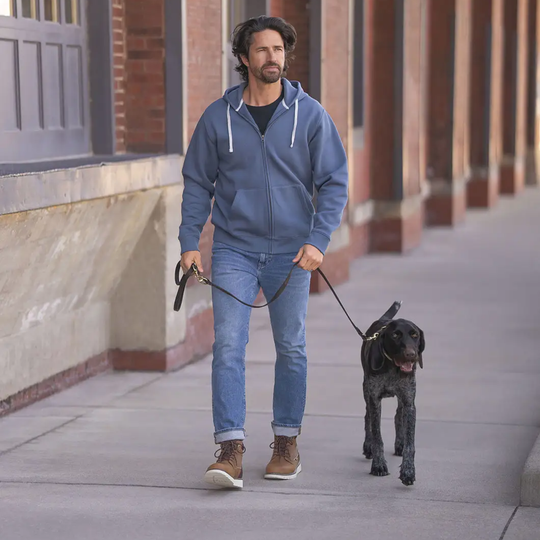 A man wearing a blue zip-up hoodie, blue jeans, and brown boots walks a black dog on a sidewalk next to a brick building.