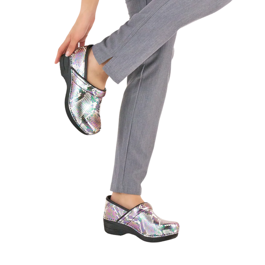 A person wearing shiny iridescent clogs with black soles, shown from the waist down against a plain white background. The person is also wearing gray pants and holding one clog with one hand.