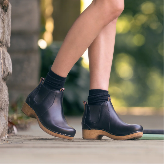 A person wearing dark navy Chelsea-style ankle boots with light brown wooden-look heels and black socks, standing outdoors on a stone ledge with a blurred green background.