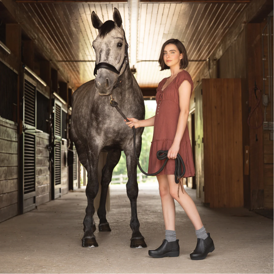 A woman in a rust-colored dress stands in a horse stable next to a dapple-gray horse, holding its reins. The stable interior has wooden stalls and a concrete floor.